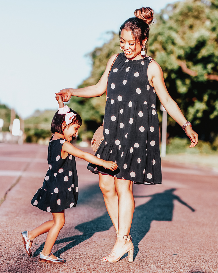 Mommy and Me Outfits featured by top US petite fashion blog Cute & Little; Image of a woman wearing LOFT Dot Tiered Swing Dress, Schutz 'Enida' Cork Sandals, pink pom earrings, Amazon Gucci dupe crossbody bag and her daughter wearing loft littles polka dot navy swing dress.