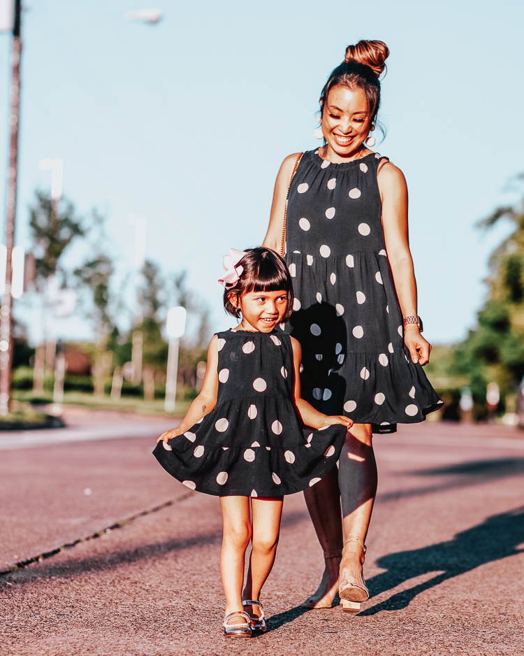 Mommy and Me Outfits featured by top US petite fashion blog Cute & Little; Image of a woman wearing LOFT Dot Tiered Swing Dress, Schutz 'Enida' Cork Sandals, pink pom earrings, Amazon Gucci dupe crossbody bag and her daughter wearing loft littles polka dot navy swing dress.