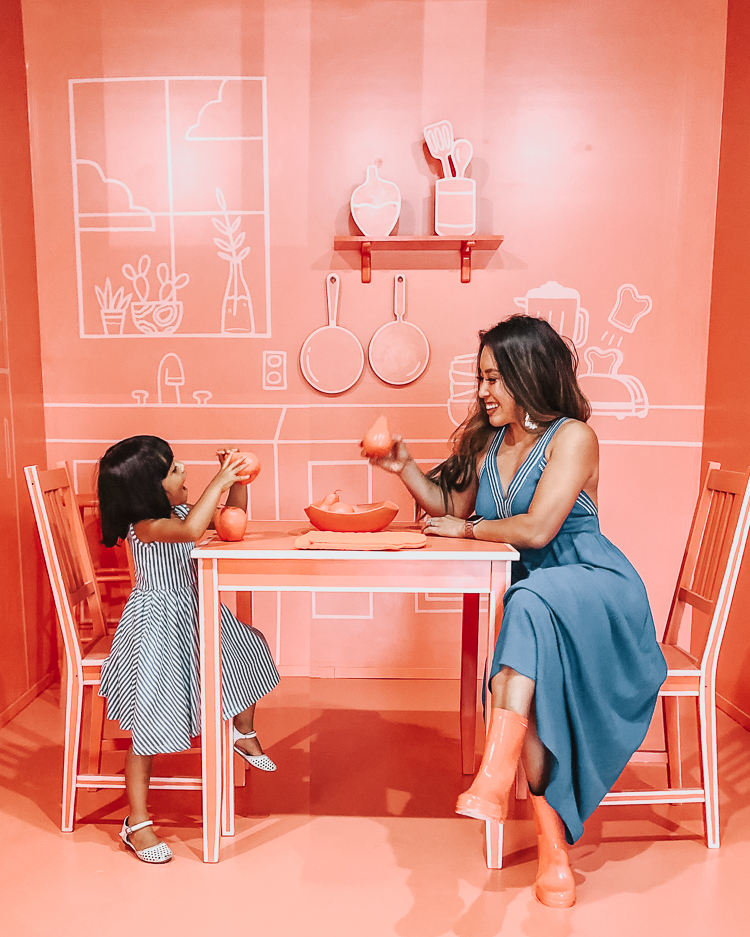 cute & little | popular dallas fashion mom blog | museum of memories instagram popup immersive art exhibit |  Take a Selfie of Your Childhood: Museum of Memories by popular Dallas blog Cute and Little: image of woman and young girl sitting at a table in a monochromatic pink room at the Museum of Memories.  The woman wearing a Gibson x Hi Sugarplum! Santorini Maxi, pink rain boots, and Panacea Shell Rope Hoops.  And the young girl is wearing an Alice & Ames Tank Ballet Dress and Nine West Kids 'Vivien' Mary Janes. 