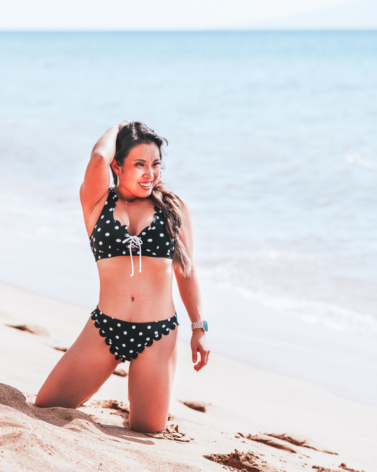 Affordable Fashion: Cute Summer Swimsuits you Need by popular Dallas blog Cute and Little: Image of a woman in Maui, Hawaii kneeling on a beach and wearing a Amazon Polka-Dot Scallop Bikini, Baublebar 'Brianna' Heart Hoops, Etsy Solitaire 15" necklace, and Fitbit 'Versa' smartwatch. 