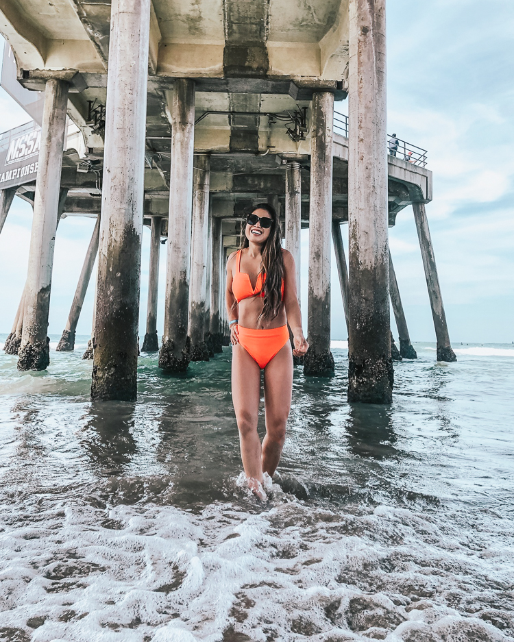 cute & little | popular dallas petite fashion blog | amazon swimsuit orange notch high waisted bikini | huntington beach california | 38 Things You May Not Know About Me by popular Dallas lifestyle blog, Cute and Little: image of a woman standing in the ocean under a pier and wearing a Amazon High Wasited Bikini Shoulder Strap 2 Piece High Cut String Swimsuit and Aerie 'Big Time' Sunnies. 