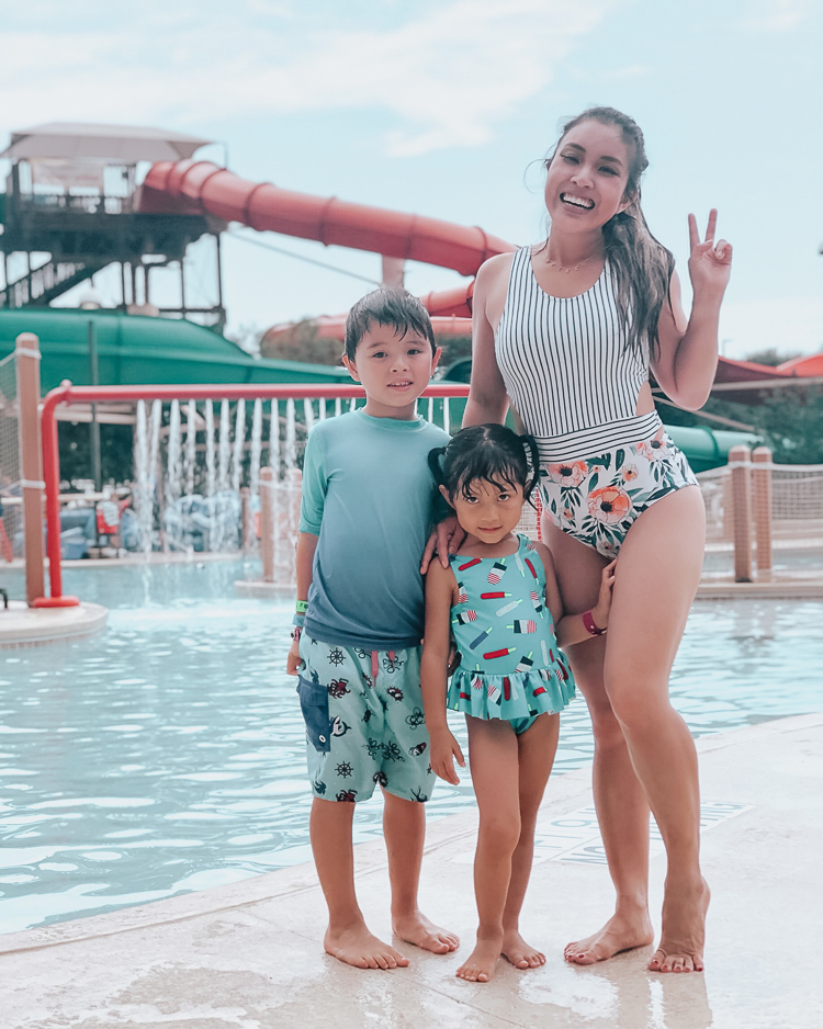 cute & little | popular dallas fashion blog | great wolf lodge grapevine water park | summer adventure vacation essentials | 4 Essentials For The Family Summer Vacation Packing List by popular Dallas petite fashion blog, Cute and Little: image of woman and two young kids standing next to a pool at a water park and wearing a floral and stripe print one piece swimsuit, blue Boys' Short Sleeve Gradient Rash Guard by Cat & Jack™, blue Boys' Shiver Me Timbers Swim Trunks by Cat & Jack™, Aqua Toddler Girls' Skirted One Piece Swimsuit by Cat & Jack™