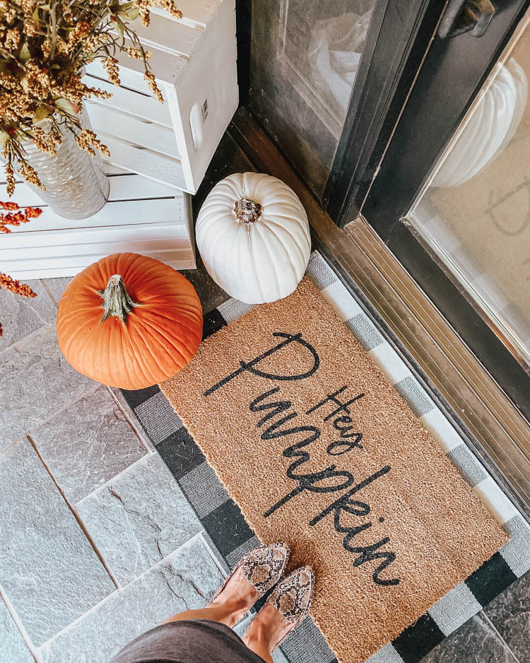 cute & little | popular dallas petite fashion lifestyle blog | how to decorate for fall on a budget | front porch fall decor doormat pumpkins | How to Get Beautiful Fall Home Decor on a Budget by popular Dallas life and style blog, Cute and Little: image of a woman holding a Trapp candle and standing out on her porch that's decorated with a Etsy Hey Pumpkin Doormat, Amazon Checkmate Cotton Buffalo Plaid Rugs Black and White Checkered Rug, Michaels Wood Crate Carry All by ArtMinds, Michaels 13" Cream Craft Pumpkin by Ashland, Michael's faux flowers, Michaels 16" Metal Whitewashed French Bucket by Ashland, and Target Metal Lantern in gold. 