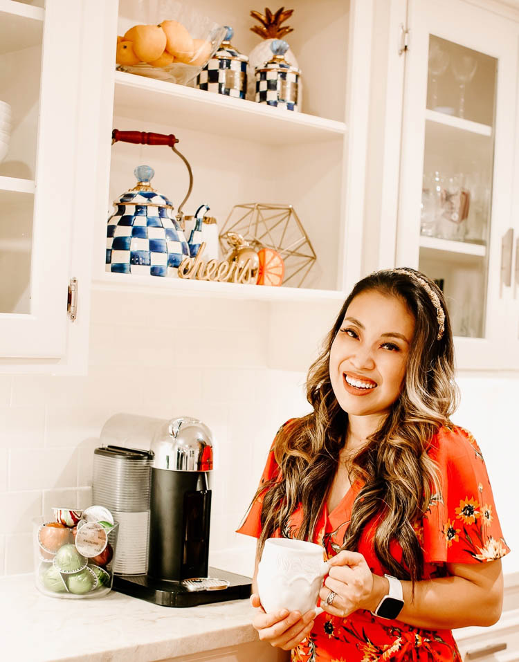cute & little | mackenzie childs royal check kitchen decor open shelving styling | A MacKenzie Childs Kitchen by popular Dallas life and style blog, Cute and Little: image of a woman wearing a red floral dress and Anthropologie Deepa Kristin Embellished Headband while holding a Mackenzie Childs Sweetbriar Mug and standing next to a Mackenzie Childs Royal Check Tea Kettle, Mackenzie Childs Royal Check Canister, and Mackenzie Childs Royal Check Canister Demi.
