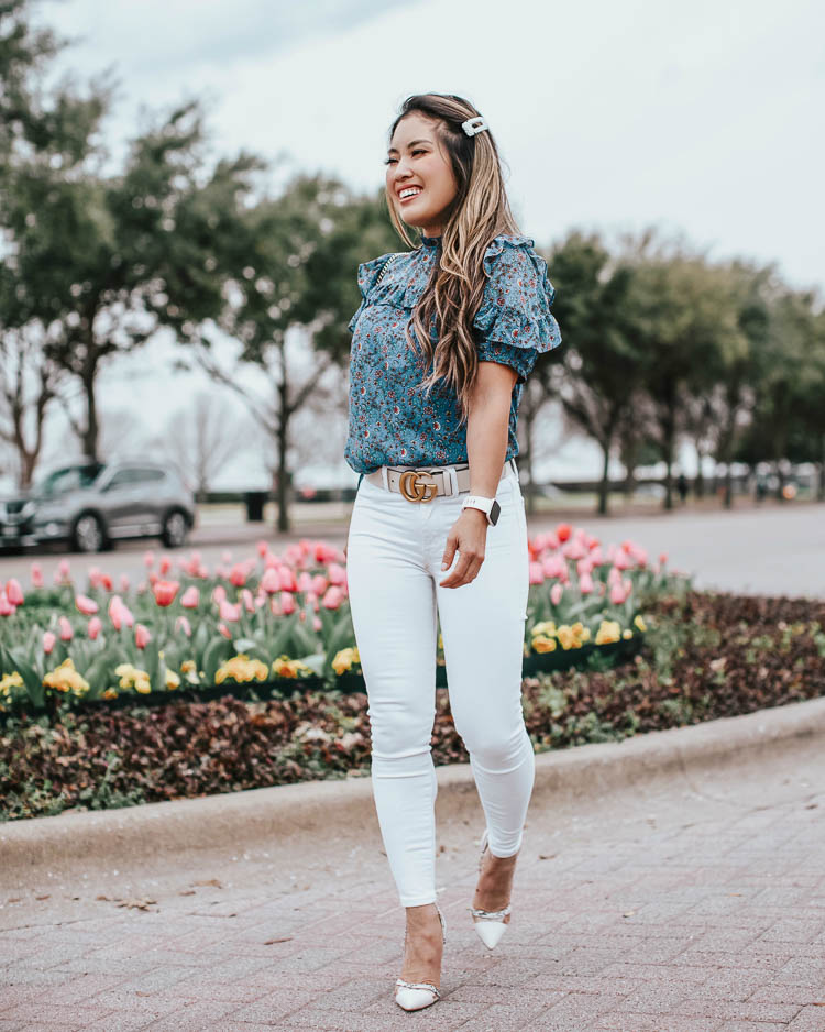 cute & little | dallas petite fashion blog | loft blue vine ruffle puff sleeve blouse, white jeans, justfab prizilla lucite pumps, white gucci belt | business casual spring work outfit | Social Distancing at Home by popular Dallas lifestyle blog, Cute and Little: image a woman walking by some tulips and wearing a Loft VINE RUFFLE PUFF SLEEVE BLOUSE, American Eagle AE NE(X)T LEVEL HIGHEST WAIST JEGGING, Just Fab Prizilla D'orsay Pump, Target Fitbit Versa Lite Smartwatch, Neiman Marcus Gucci GG Marmont Chevron Quilted Leather Flap Wallet on a Chain, and Sephora HUDA BEAUTY Liquid Matte Lipstick.