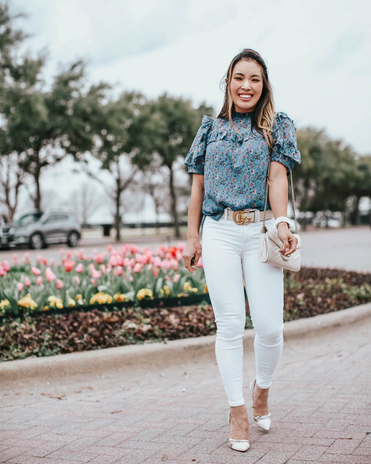 cute & little | dallas petite fashion blog | loft blue vine ruffle puff sleeve blouse, white jeans, justfab prizilla lucite pumps, white gucci belt | business casual spring work outfit | Social Distancing at Home by popular Dallas lifestyle blog, Cute and Little: image a woman walking by some tulips and wearing a Loft VINE RUFFLE PUFF SLEEVE BLOUSE, American Eagle AE NE(X)T LEVEL HIGHEST WAIST JEGGING, Just Fab Prizilla D'orsay Pump, Target Fitbit Versa Lite Smartwatch, Neiman Marcus Gucci GG Marmont Chevron Quilted Leather Flap Wallet on a Chain, and Sephora HUDA BEAUTY Liquid Matte Lipstick.