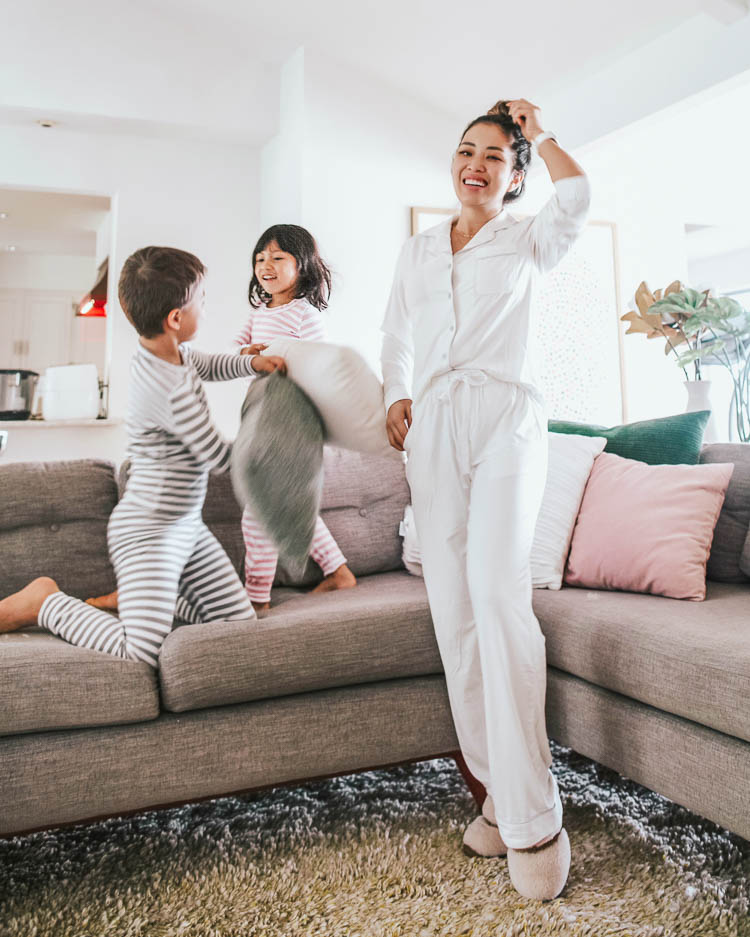 cute & little | dallas fashion mom blogger | mothers day 2020 gift guide ideas | Mother's Day Gift Ideas by popular Dallas life and style blog, Cute and Little: image of a woman wearing a Cozy Earth pajama set, Amazon UGG Women's Dalla Slipper, and standing next to her kids who are hitting each other with pillows on their sectional couch and wearing Monica + Andy pajamas. 