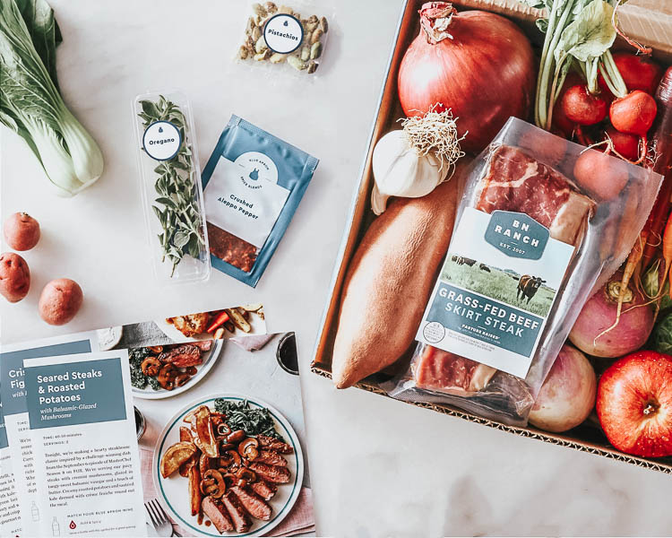 comparing 5 different home meal delivery kits | best meal delivery kit | blue apron review | Meal Delivery Service Reviews by popular Dallas lifestyle blog, Cute and Little: image of some Blue Apron recipes, a box full of fruit, vegetables, BN Ranch grass-fed beef skirt steak, and some Bok Choy and herbs on a marble counter top. 