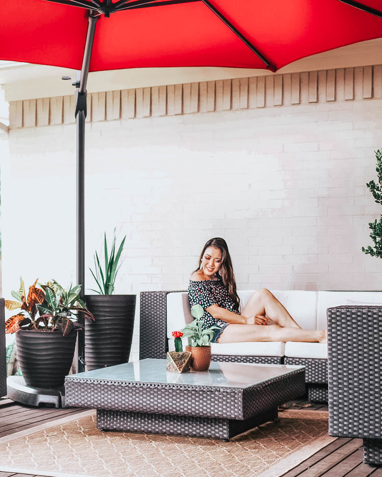 cute & little | dallas fashion lifestyle blogger | backyard patio | Backyard Patio by popular Dallas life and style blog, Cute and Little: image of a woman sitting on a outdoor sectional next to a woven indoor/outdoor rug under a Lowe's Simply Shade Red Offset 11-ft Auto-tilt Octagon Patio Umbrella.