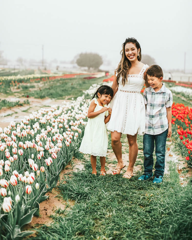 cute & little | dallas family fashion blog | free people verona white dress | texas tulips farm | spring summer family picture outfits | Tulip Farm by popular Dallas lifestyle blog, Cute and Little: image of a mom and her two young children standing next to a row of tulips at the Texas Tulip Farm and wearing a Free People 'Verona', Marc Fisher 'Arena' sandals, Baublebar Lady Button stud earrings, yellow ruffle dress, strappy sandals, plaid shirt, pull-on jeans, and sneakers.