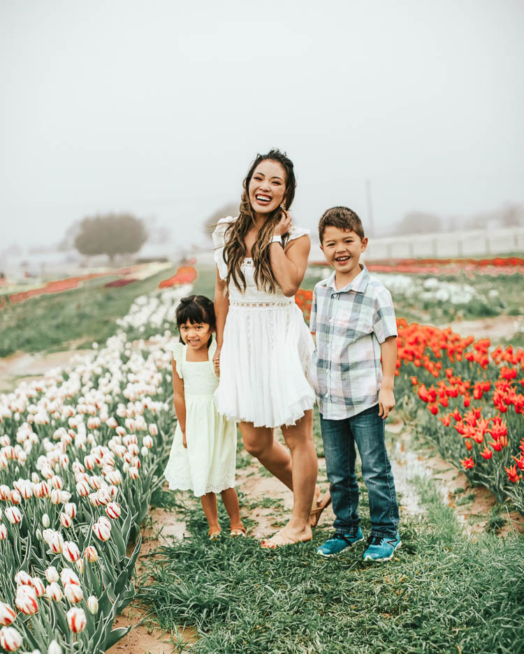 cute & little | dallas family fashion blog | free people verona white dress | texas tulips farm | spring summer family picture outfits | Tulip Farm by popular Dallas lifestyle blog, Cute and Little: image of a mom and her two young children standing next to a row of yellow tulips at the Texas Tulip Farm and wearing a Free People 'Verona', Marc Fisher 'Arena' sandals, Baublebar Lady Button stud earrings, yellow ruffle dress, strappy sandals, plaid shirt, pull-on jeans, and sneakers.