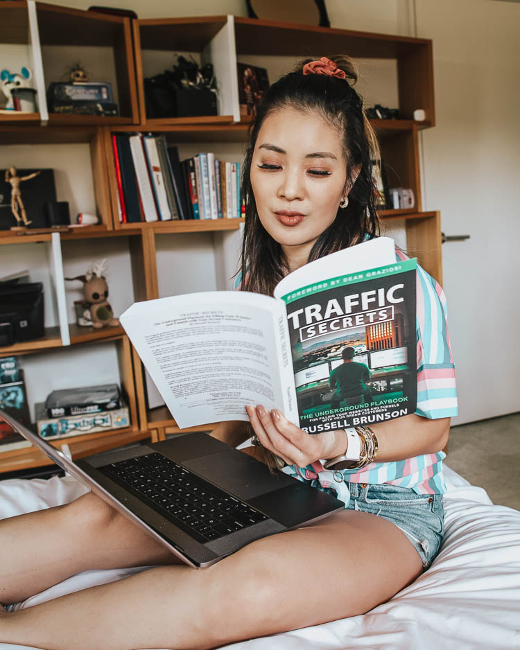 cute & little | dallas petite fashion lifestyle blog | tips build boost blog traffic | How to Increase Blog Traffic by popular Dallas lifestyle blog, Cute and Little: image of a woman wearing pajamas and sitting on her bed with a Traffic Secrets book in on hand and a laptop in her lap. 
