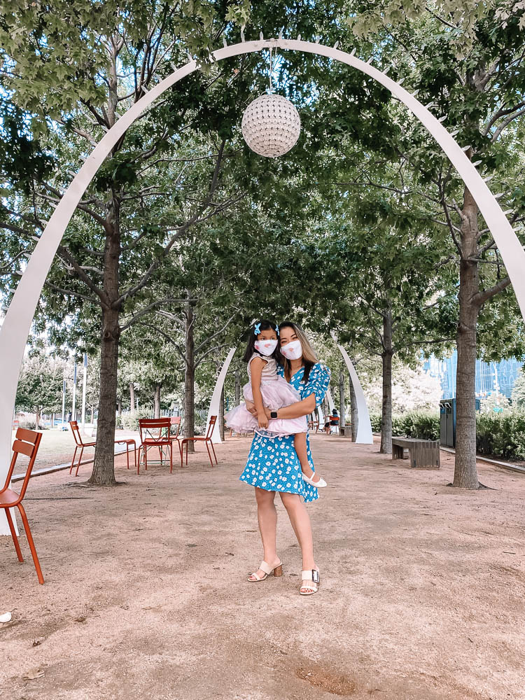 cute & little | dallas family mom travel blog | ritz-carlton dallas staycation | klyde warren park | Family Staycation by popular Dallas lifestyle blog, Cute and Little: image of a mom holding her daughter at the Klyde Warren Park and wearing a Chicwish Daisy Wrap Midi Dress, Swaddle Designs watercolor floral face mask, Amazon find. Women's Large Buckle Block Heel Sandal, Pippa & Julie Tanya Holographic Dress, Target Glitter Ballet Flats, Swaddle Designs kids' watercolor floral face mask, and blue Amazon hair bows.  