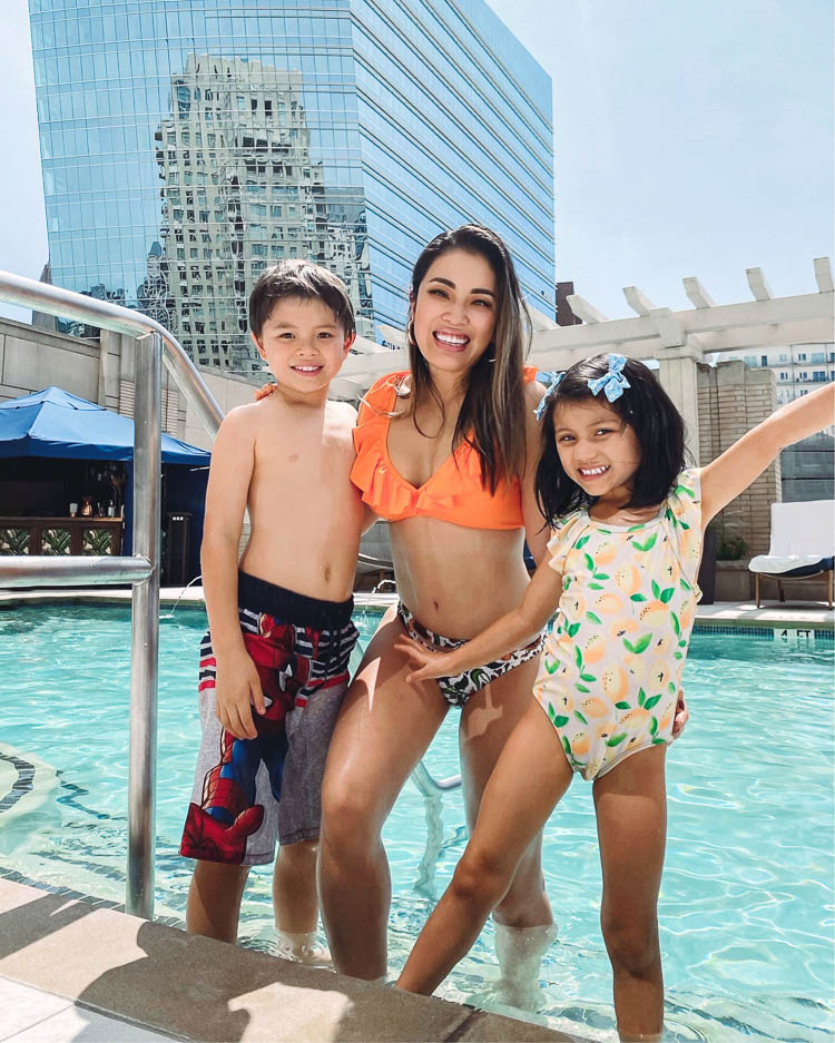 cute & little | dallas family mom travel blog | ritz-carlton dallas staycation | Family Staycation by popular Dallas lifestyle blog, Cute and Little: image of a mom and her son and daughter standing in a swimming pool and wearing a Amazon CUPSHE Women's Orange Floral Bottom Ruffle Hook Closure Bikini Set, Target Boys' Spider-Man Print Swim Trunks, and Target Toddler Girls' Strawberry Flutter Sleeve One Piece Swimsuit.