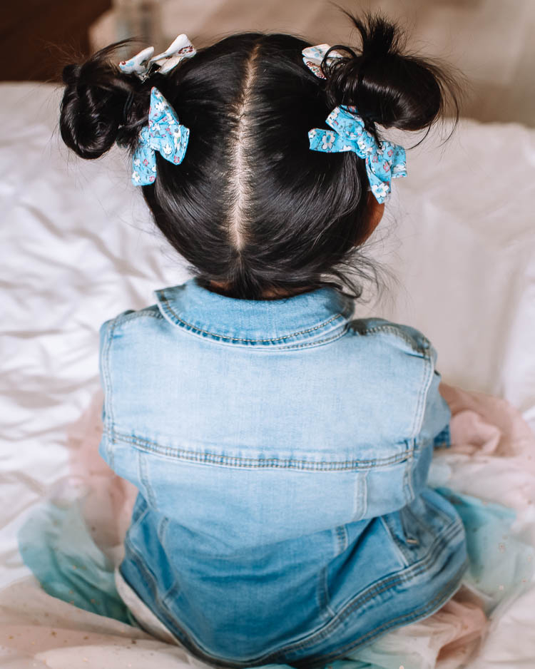 cute & little | dallas mom blog | messy pigtail space buns hairstyle school tutorial | Pigtail Buns by popular Dallas beauty blog, Cute and Little: image of a little girl with a pigtail buns hairstyle and wearing a Target Girls' Jean Jacket, Pippa and Julie Odette Star Rainbow Dress, and Amazon CellElection Hair Bows Clips while sitting on a bed. 
