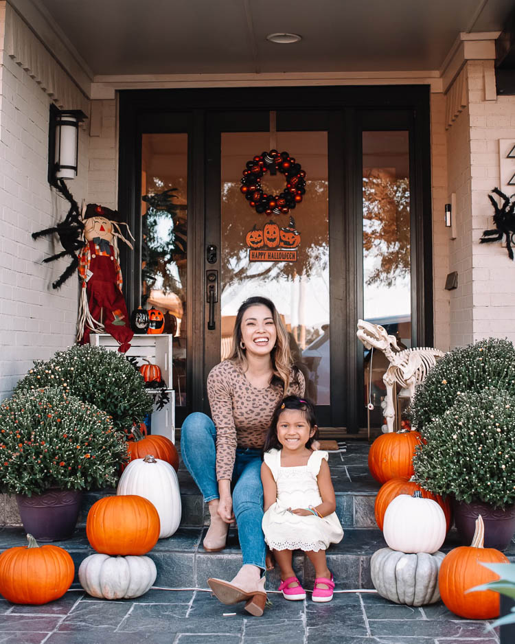cute & little | dallas mom petite fashion blog | halloween front porch fall decor | walmart plus membership review |Walmart Halloween Decor by popular Dallas lifestyle blog, Cute and Little: image of a woman and her daughter sitting on their front porch decorated with a Walmart pumpkin wreath, pumpkin trio sign scarecrow, similar crates | similar light-up pumpkin similar 'hey pumpkin' doormat, buffalo check doormat giant spider, dog skeleton, and pumpkin pathway lights.