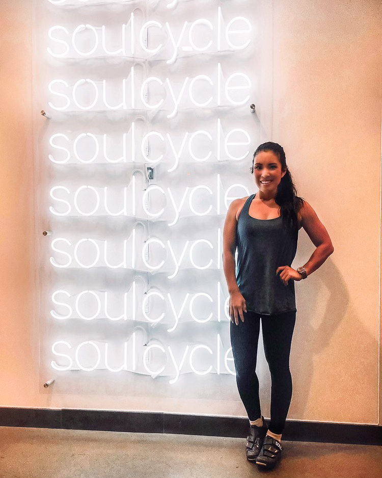 cute & little | dallas petite fashion fitness blog | soulcycle review |SoulCycle At Home Bike by popular Dallas lifestyle blog, Cute and Little: image of a woman wearing a black tank and black leggings and standing next to a neon SoulCycle sign. 