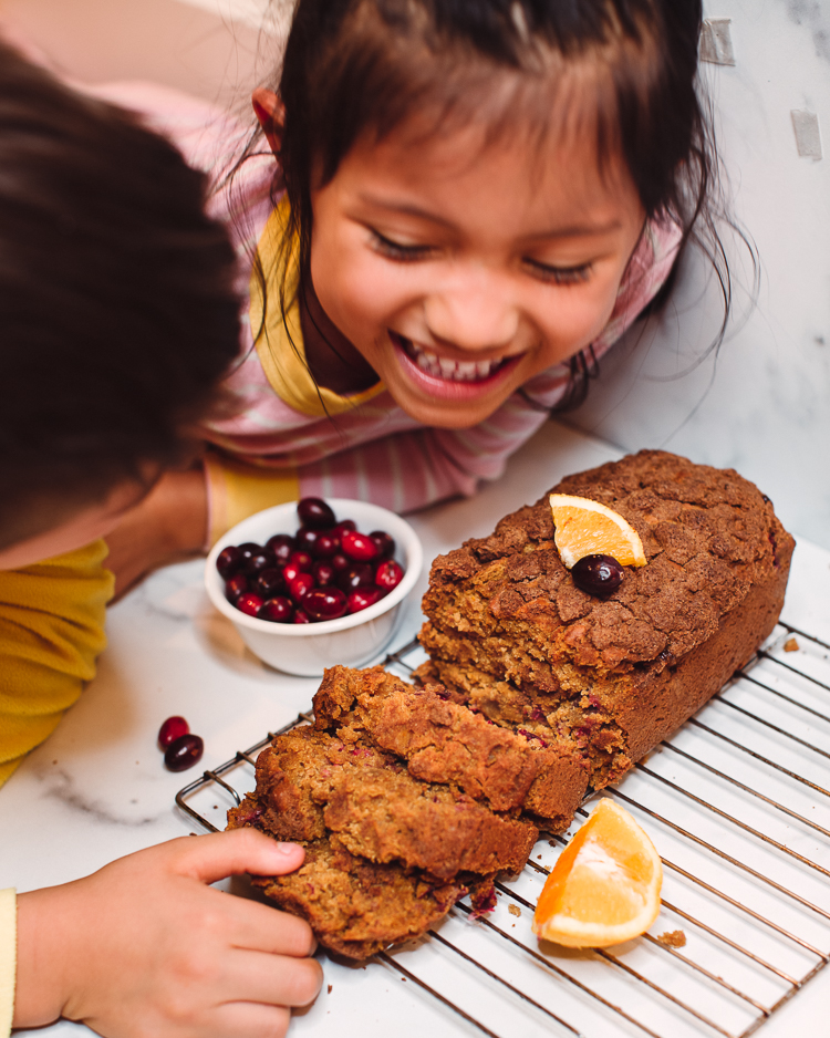 cute & little | dallas petite mom lifestyle blog | thanksgiving christmas holiday dessert recipe | cranberry orange quick bread |Cranberry Orange Quick Bread Recipe by popular Dallas lifestyle blog, Cute and Little: image of two young children leaning over a cranberry orange quick bread loaf that's on a cooling rack. 