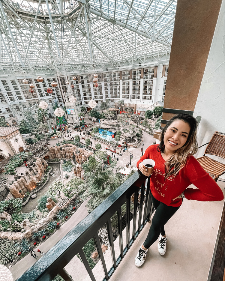 cute & little | dallas family mom blog | gaylord texas lone star christmas review |Gaylord Texan by popular Dallas lifestyle blog, Cute and Little: image of a woman wearing a read sweater, black leggings and Golden Goose sneakers while standing on hotel room balcony at the Gaylord Texan. 