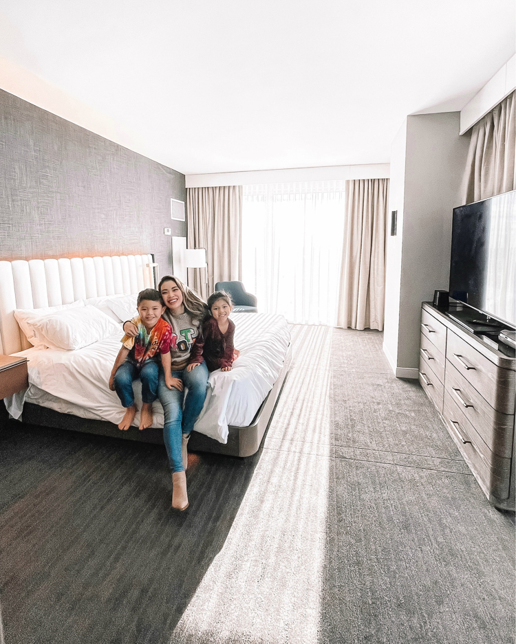 cute & little | dallas mom lifestyle blog | things to do frisco holidays 2020 | hyatt regency frisco review |Things to do in Frisco TX by popular Dallas travel blog, Cute and Little: image of a mom and her two kids sitting on a bed in Hyatt Regency room. 