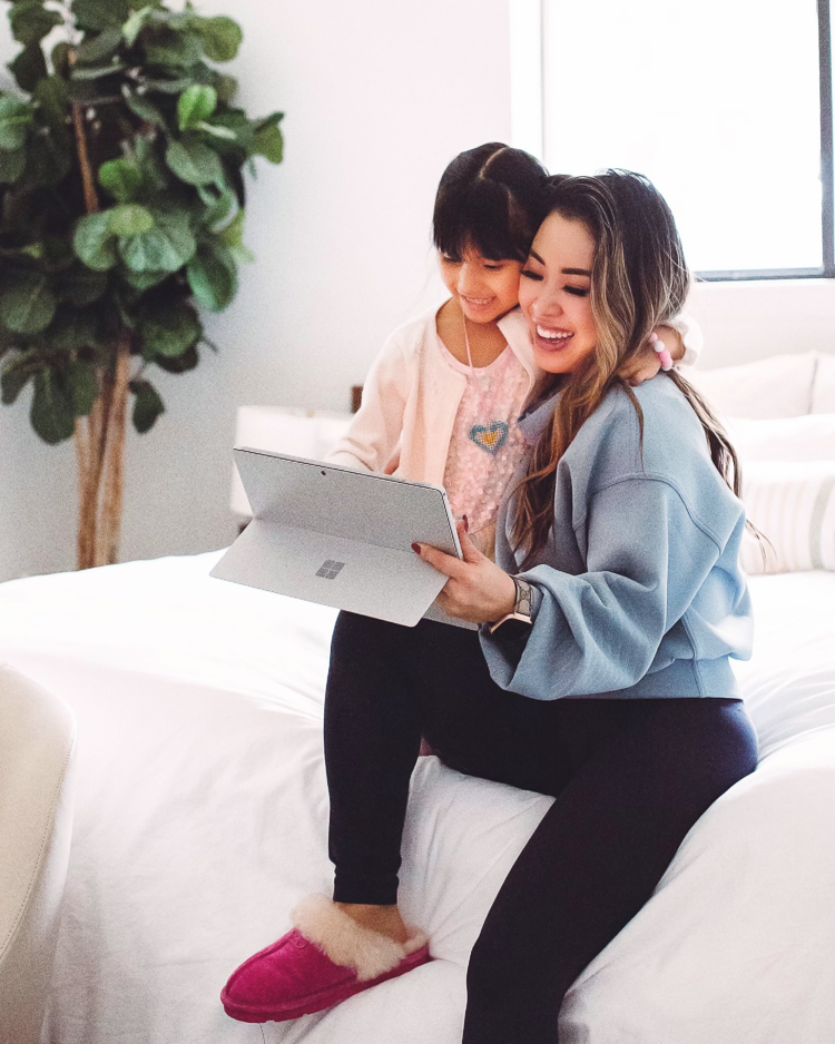 cute & little | dallas petite fashion blog | lululemon full flourish pullover, microsoft surface pro 7 | imposter syndrome woman tech | How to Overcome Imposter Syndrome by popular Dallas lifestyle blog, Cute and Little: image of a mom wearing a Lululemon fleece pullover, black leggings, and pink fur lined Ugg slippers and sitting next to her daughter on a bed with white bedding and looking at a tablet. 
