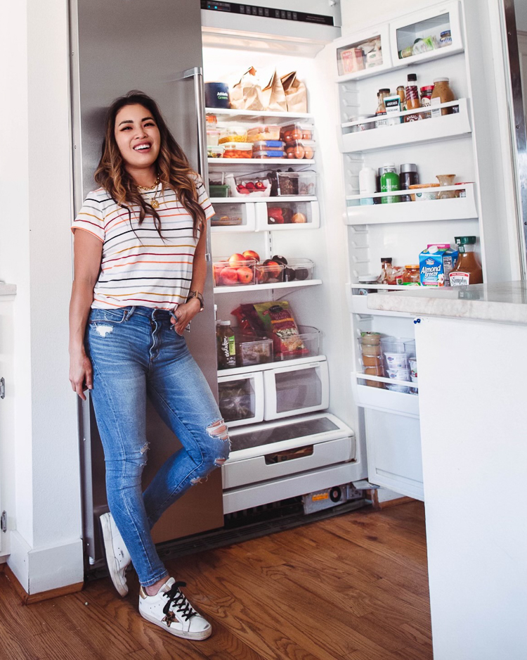 cute & little | dallas mom lifestyle blog | fridge organization tips and must-haves from walmart | Walmart Fridge Organization by popular Dallas lifestyle blog, Cute and Little: image of a woman wearing a rainbow stripe t-shirt, distressed jeans, and Golden Goose sneakers while leaning against her open fridge. 