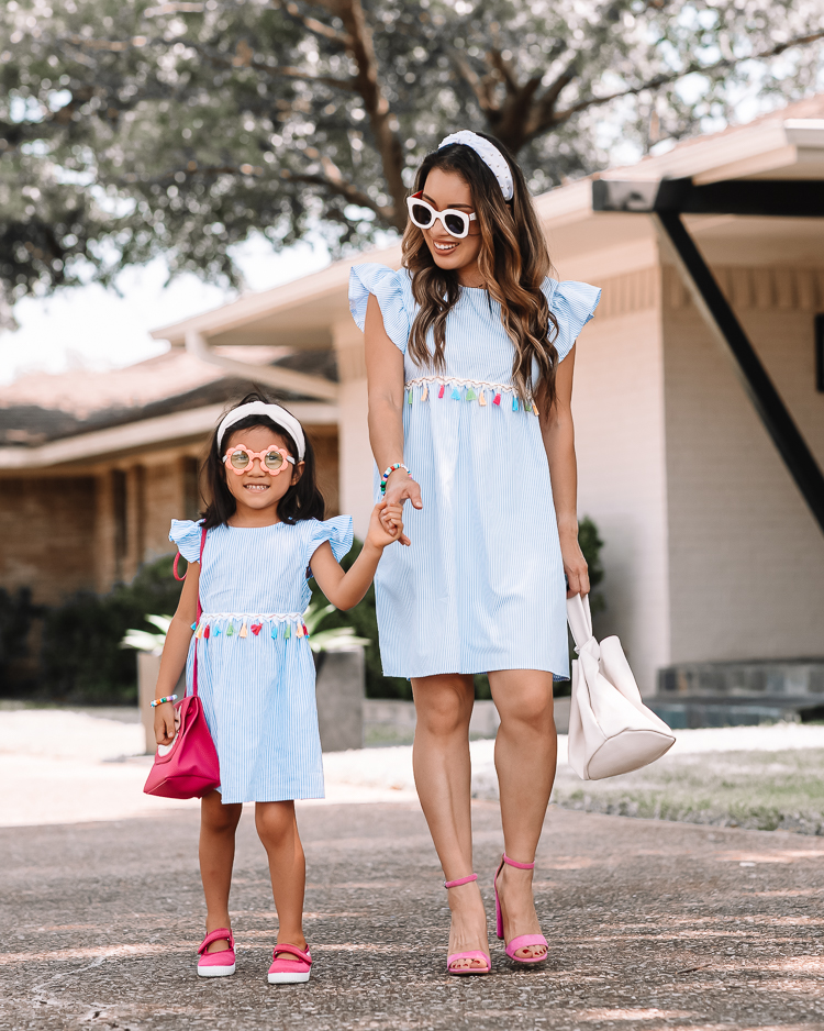 cute & little | dallas mom petite fashion blog | mom daughter matching spring summer dress outfits | Mommy and Me Outfits by popular Dallas petite fashion blog, Cute and Little: image of a mom and her daughter standing outside and wearing matching blue and white stripe tassel dresses, white knot headbands, sunglasses, and pink shoes. 