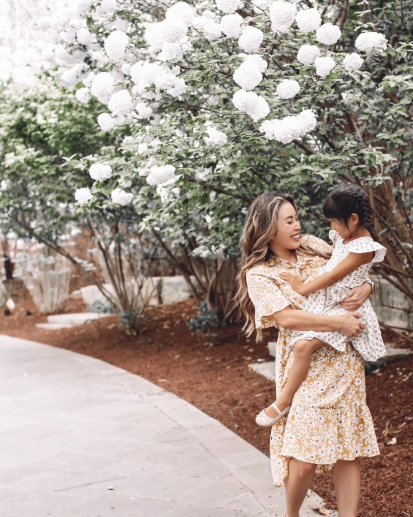 cute & little | dallas petite fashion blog | amazon spring summer dresses mom daughter wedding guest graduation vacation photoshoot | Spring Dresses From Amazon by popular Dallas petite fashion blog, Cute and Little: image of a mom holding her daughter under a tall white flowering bush and wearing floral print spring dresses. 
