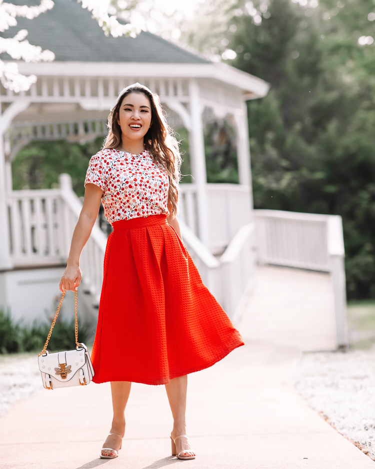 cute & little | dallas mom petite fashion blog | cherry tee, red retro midi skirt | mothers day gift guide 2021 | Mother's Day Gifts by popular Dallas life and style blog, Cute and Little: image of a woman standing in front of a white gazebo and wearing a LOFT Cherry Crew Tee, red pleated skirt, Dolce Vita 'Noles', white knot headband, and Fitbit 'Versa 2'  w/ leopard Spark*l Band
