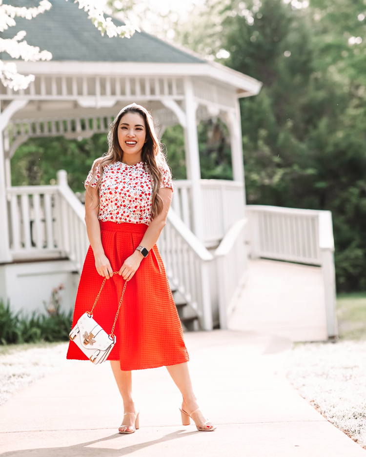 cute & little | dallas mom petite fashion blog | cherry tee, red retro midi skirt | mothers day gift guide 2021 |Mother's Day Gifts by popular Dallas life and style blog, Cute and Little: image of a woman standing in front of a white gazebo and wearing a LOFT Cherry Crew Tee, red pleated skirt, Dolce Vita 'Noles', white knot headband, and Fitbit 'Versa 2'  w/ leopard Spark*l Band