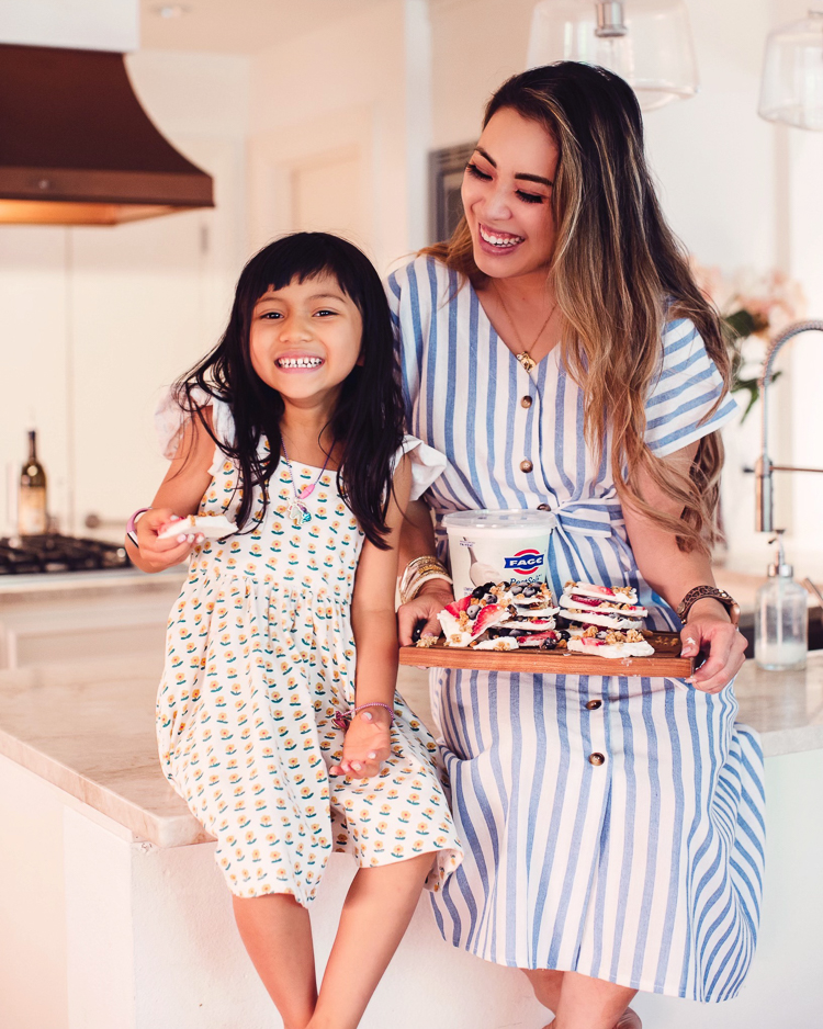 cute & little | dallas mom blog | summertime healthy snack | yogurt bark with berries | Yogurt Bark With Berries by popular Dallas lifestyle blog, Cute and Little: image of a woman holding a wooden board with yogurt bark and Fage yogurt while sitting on the kitchen counter next to her daughter. 