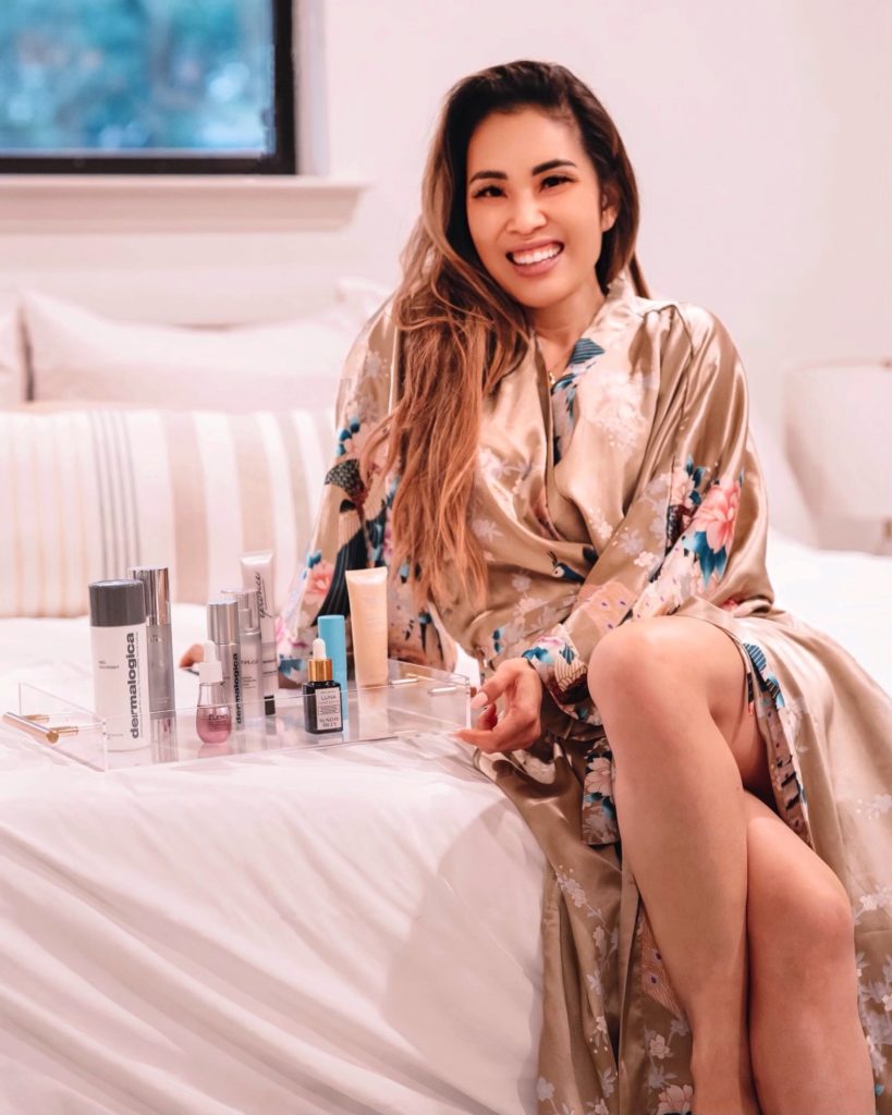 cute & little | dallas beauty blogger | dermstore anniversary sale: must-have beauty | skincare hair | Dermstore Anniversary Sale by popular Dallas beauty blog, Cute and Little: image of a woman wearing a silk floral print robe and sitting on a bed next to an acrylic tray filled with beauty products. 